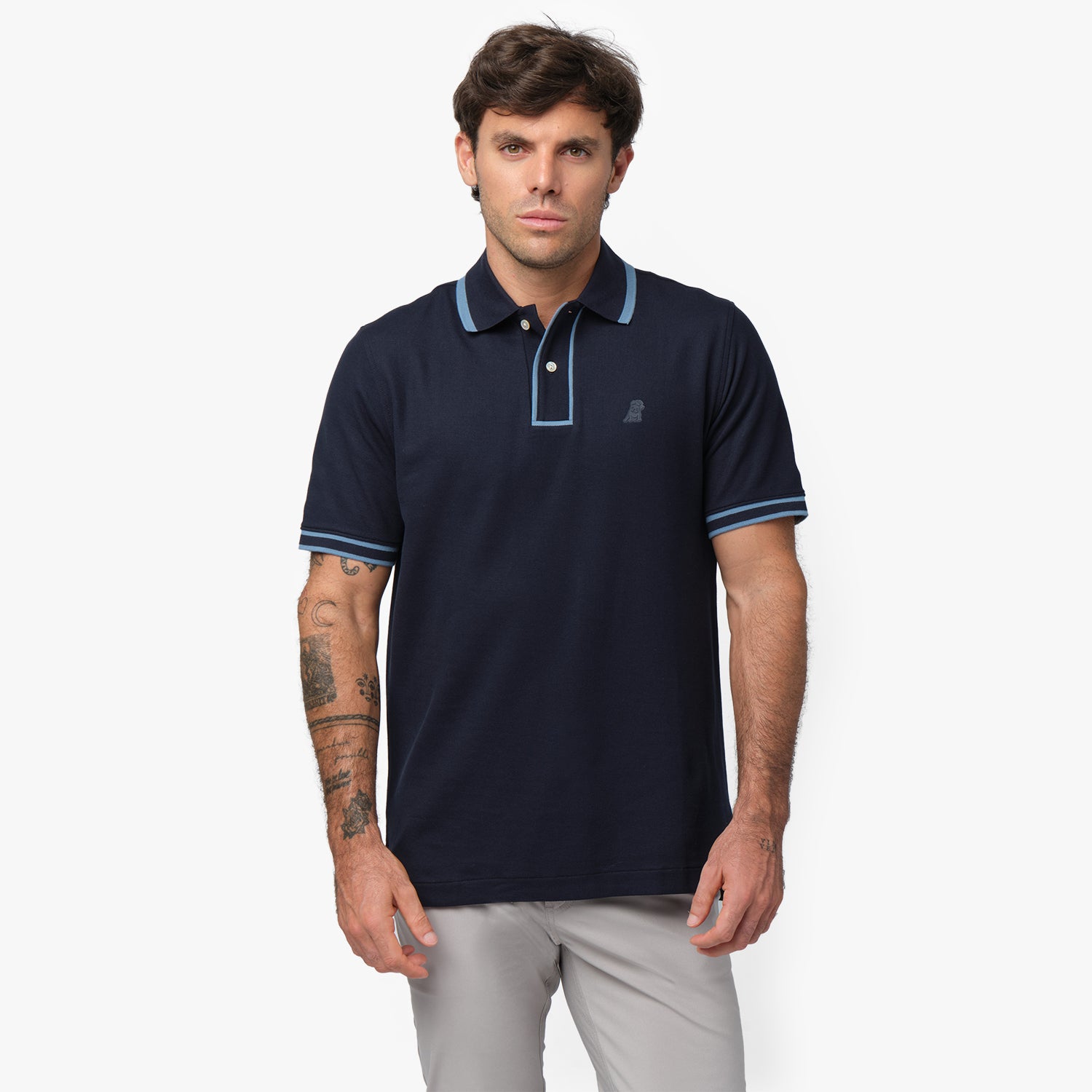 Male model wearing a navy blue short-sleeve polo shirt with contrasting light blue details on the collar, button placket, and sleeve cuffs. The polo features a two-button design and a subtle embroidered logo on the chest. Paired with light gray trousers for a refined, casual look