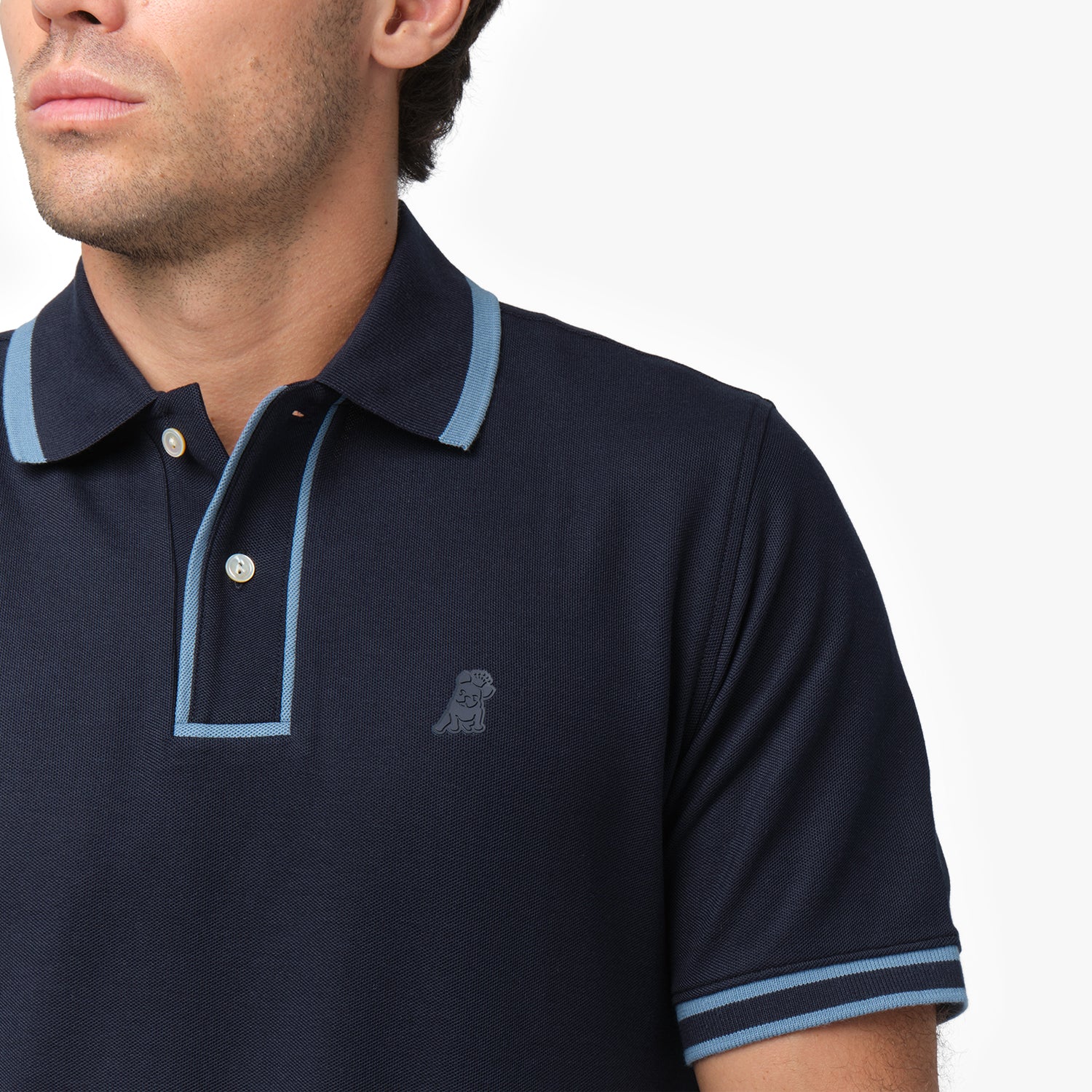 Close-up of a male model wearing a navy blue short-sleeve polo shirt with contrasting light blue details on the collar, button placket, and sleeve cuffs. The polo features a two-button design and a subtle embroidered dog logo on the chest, blending seamlessly with the fabric