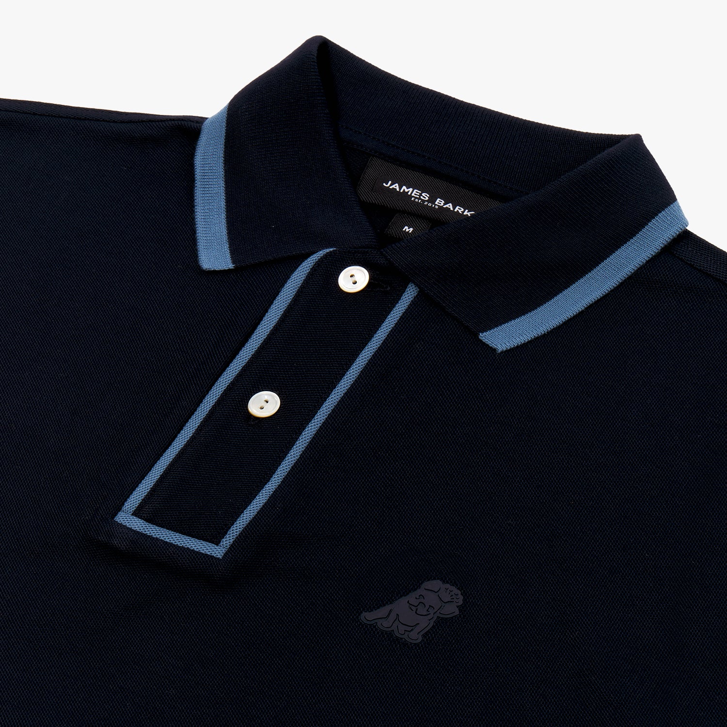 Close-up of a navy blue polo shirt featuring a ribbed collar with contrasting light blue trim. The two-button placket is outlined with matching light blue piping, adding a refined touch. The fabric has a textured pique finish, and an embroidered tonal dog logo is subtly placed on the left chest. The brand label "James Bark" is visible inside the collar.