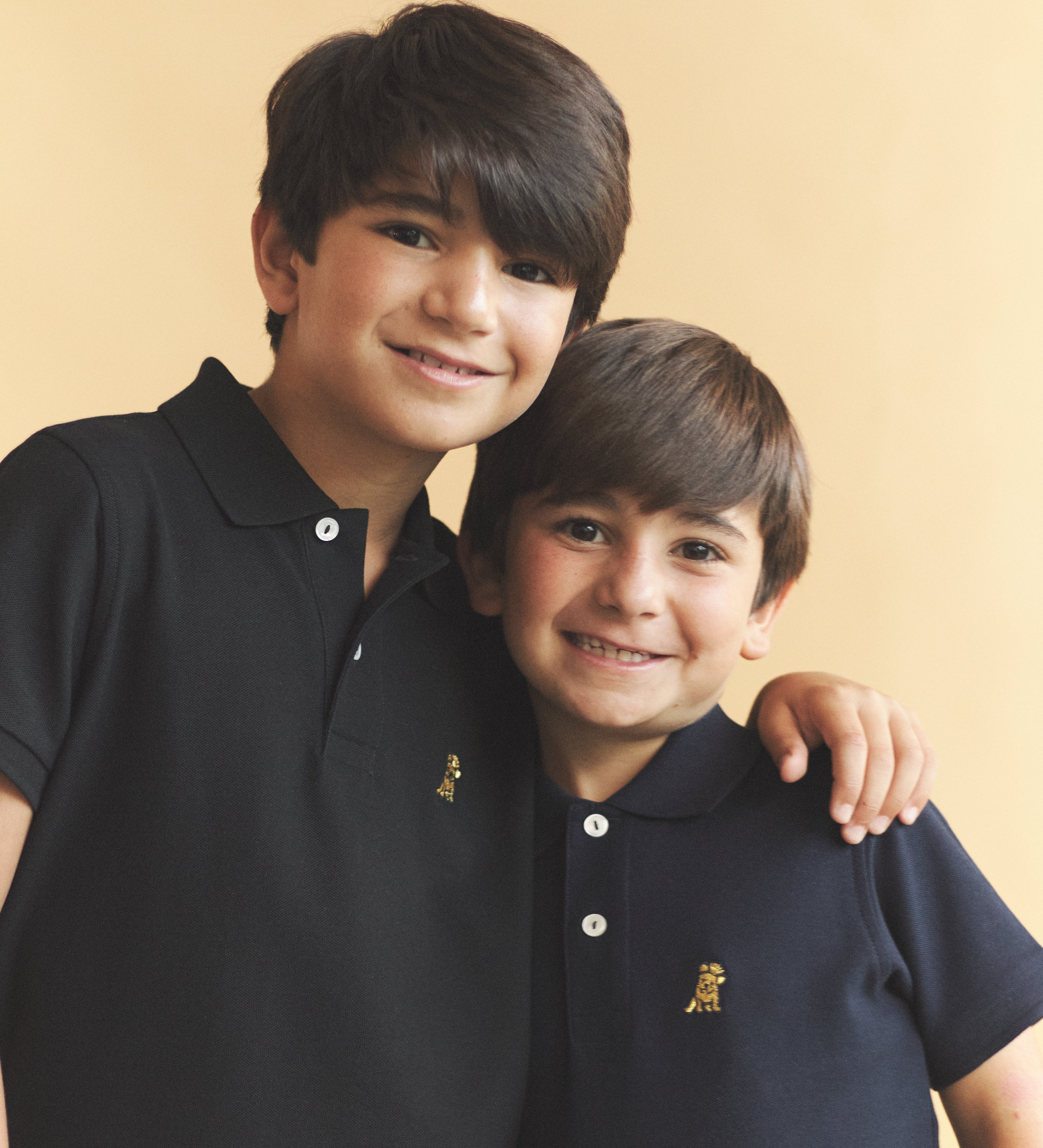 Two children wearing matching black polo shirts with a logo against a beige background