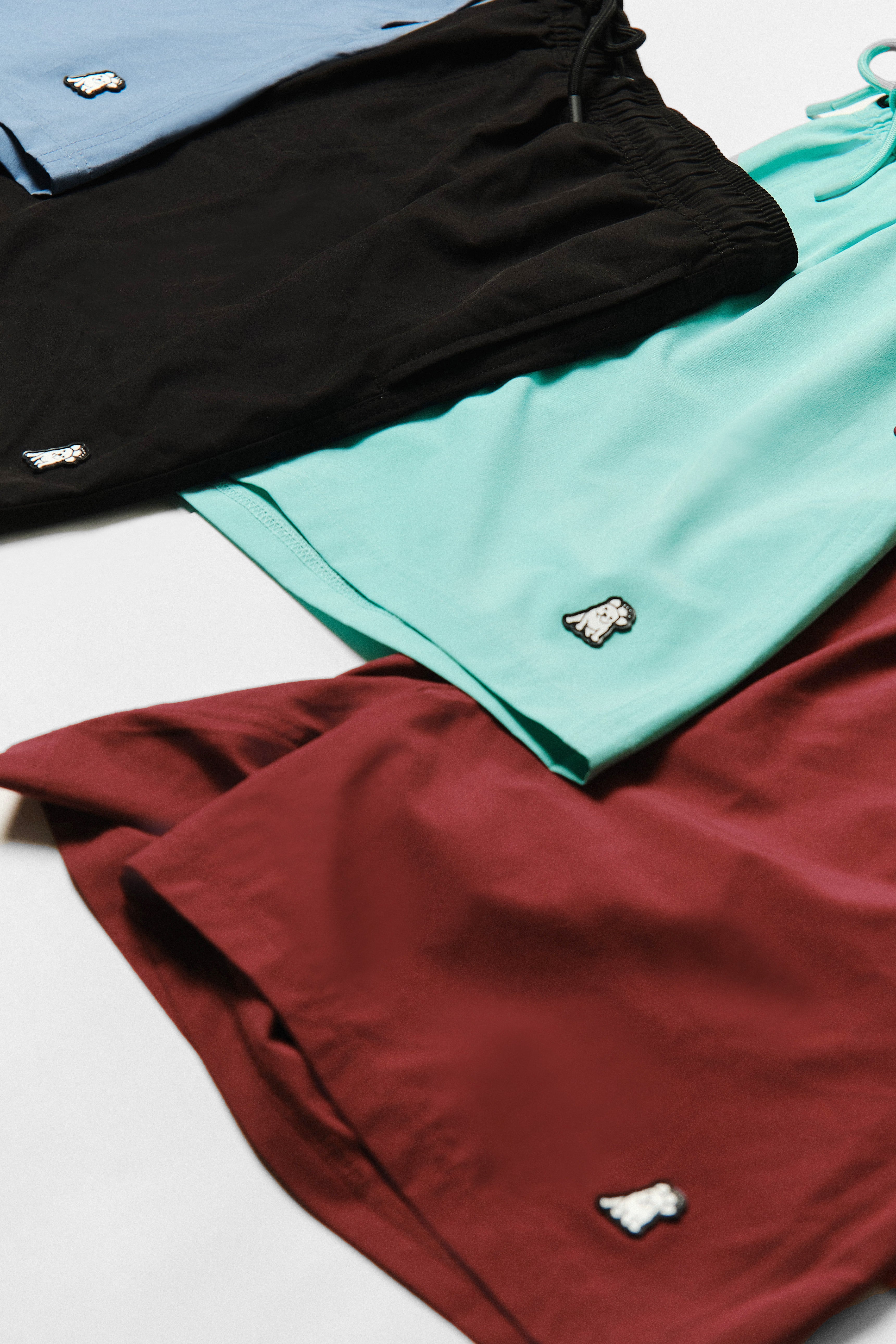 A neatly arranged collection of colorful shorts in blue, black, mint green, and burgundy. Each pair of shorts features a small embroidered dog logo near the hem. The shorts are laid out on a flat surface, highlighting their soft fabric and casual style.