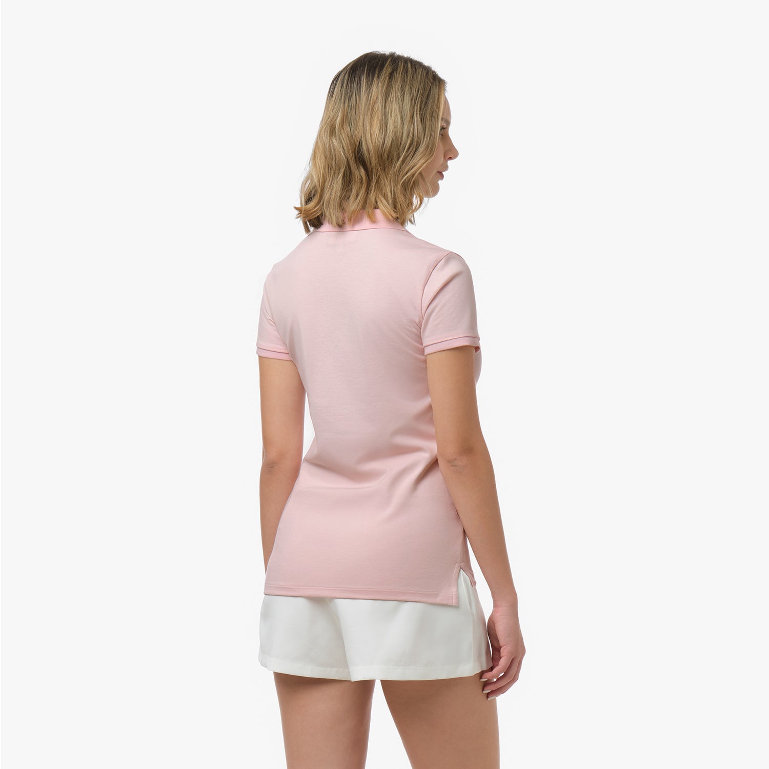 Back view of a woman wearing a short-sleeved light pink polo shirt with side slits and white shorts against a white background