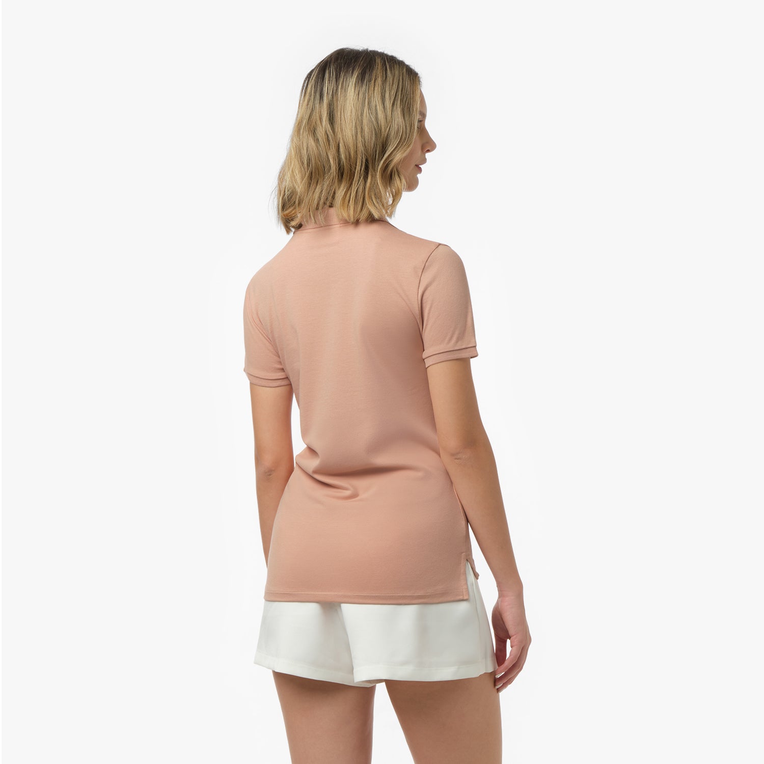 Back view of a woman wearing a short-sleeved peach polo shirt with side slits and white shorts against a white background