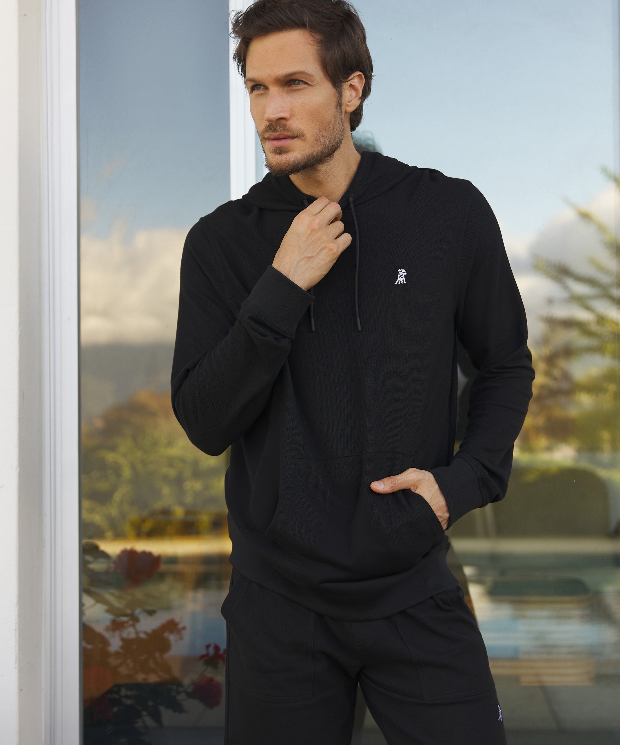 A man wearing a black hoodie and black shorts stands next to a large window, looking away from the camera