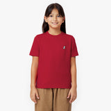 Front view of a young girl smiling, wearing a red t-shirt with a small embroidered logo and brown pants, against a white background