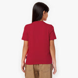Back view of a young girl wearing a red t-shirt with a small tag on the side seam and brown pants, against a white background