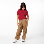 A young girl stands with her hands in the pockets of her brown pants, looking down, wearing a red t-shirt with a small logo and white sneakers, against a white background