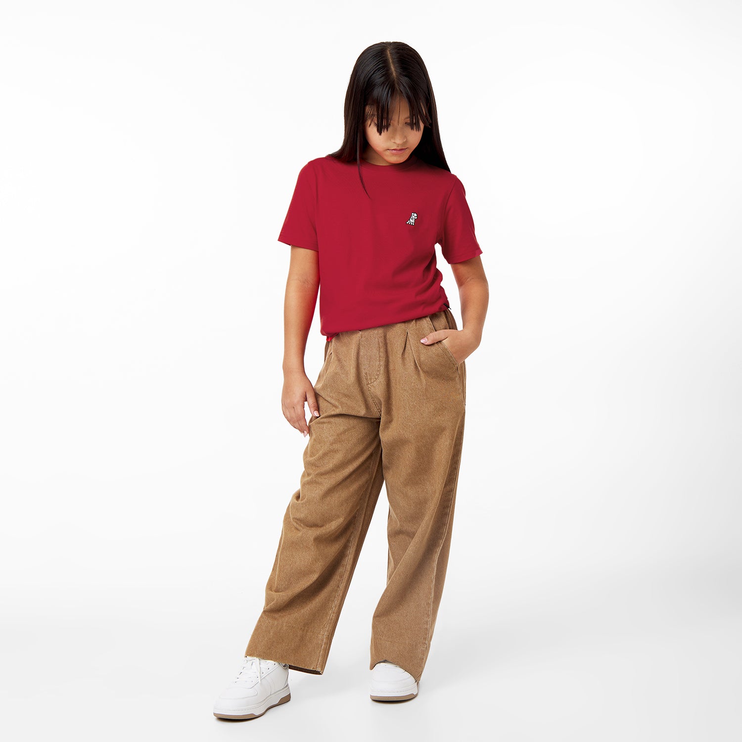 A young girl stands with her hands in the pockets of her brown pants, looking down, wearing a red t-shirt with a small logo and white sneakers, against a white background