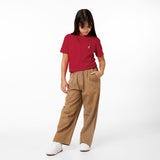 A young girl stands with her hands in the pockets of her brown pants, looking down, wearing a red t-shirt with a small logo and white sneakers, against a white background