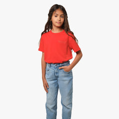 A young girl wearing a red T-shirt with a subtle graphic logo on the chest, paired with blue jeans, standing with her hand in her pocket and smiling.