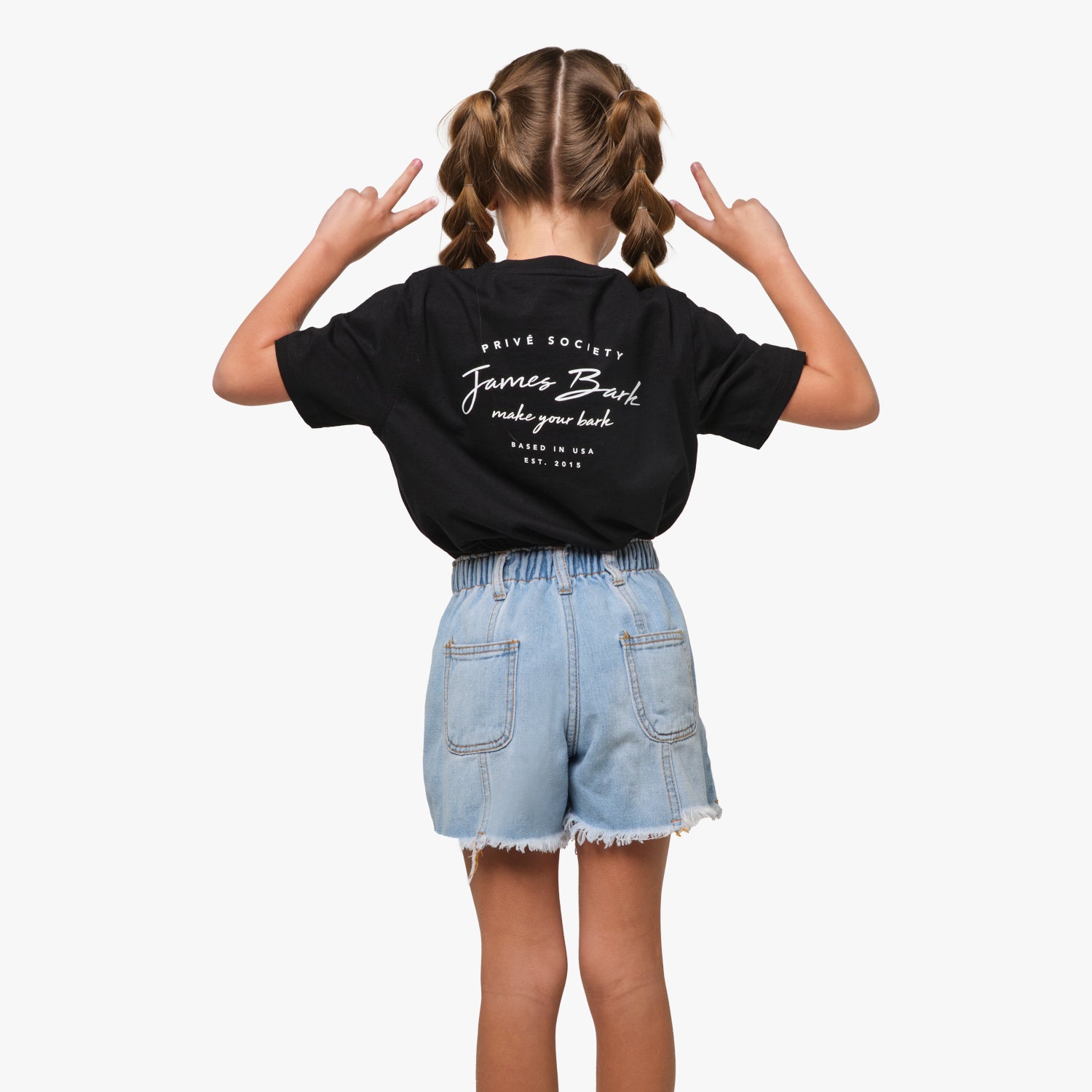 Rear view of a young girl wearing a black T-shirt with a cursive text graphic on the back, paired with denim shorts, playfully posing with peace signs.