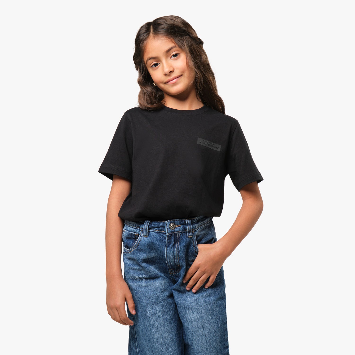 A young girl wearing a black T-shirt with a subtle graphic logo on the chest, paired with blue jeans, standing with her hand in her pocket and smiling.