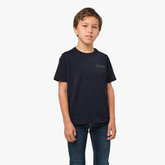 A young boy wearing a navy blue T-shirt with a subtle graphic logo on the chest, paired with jeans, standing with his hands at his sides.