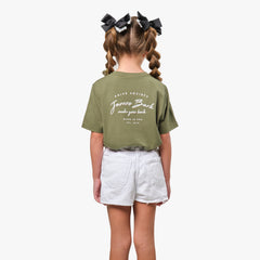 Rear view of a young girl wearing an olive green T-shirt with a cursive text graphic on the back, paired with white shorts and bow accessories in her hair.