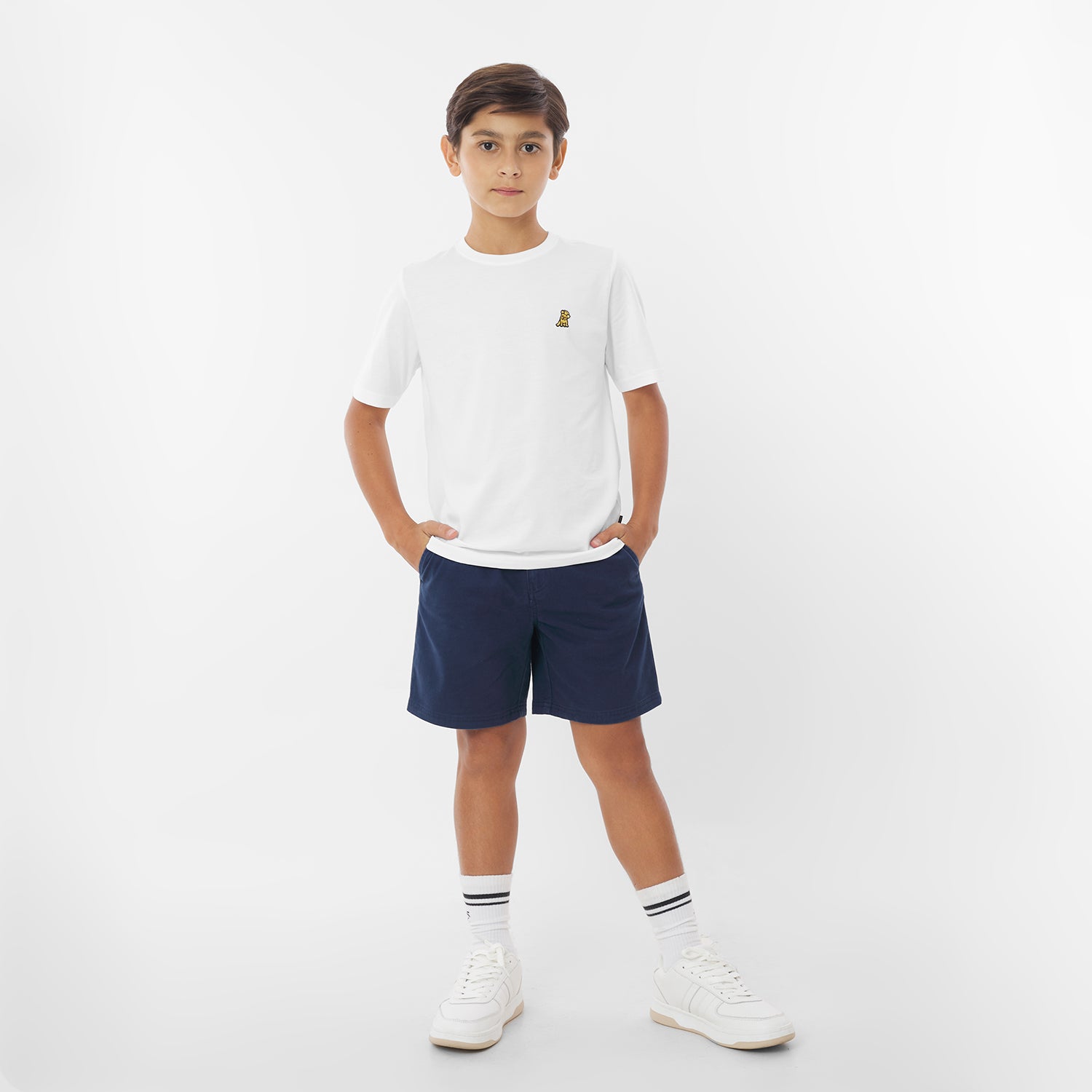 A young boy wearing a white t-shirt with a yellow logo, navy blue shorts, white socks, and white sneakers stands against a white background with his hands in his pockets