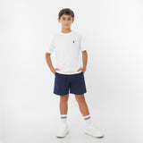 A young boy wearing a white t-shirt with a yellow logo, navy blue shorts, white socks, and white sneakers stands against a white background with his hands in his pockets