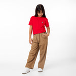 A young girl with long dark hair is wearing a red t-shirt with a small embroidered dog logo on the left chest. She pairs it with loose-fitting brown corduroy pants and white sneakers. She has one hand in her pocket and is looking down, standing against a clean white background.