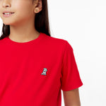 Close-up of a young girl wearing a vibrant red t-shirt with a small embroidered dog logo on the left chest. The fabric appears soft and slightly ribbed. She has dark brown hair and is wearing simple gold stud earrings. The image captures part of her face, showing a subtle smile against a clean white background.