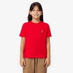 A young girl with long, dark brown hair and a bright smile wears a vibrant red short-sleeve t-shirt with a small embroidered dog logo on the left chest. She pairs it with loose-fitting brown pants. She stands against a clean white background, looking directly at the camera with a relaxed and happy expression