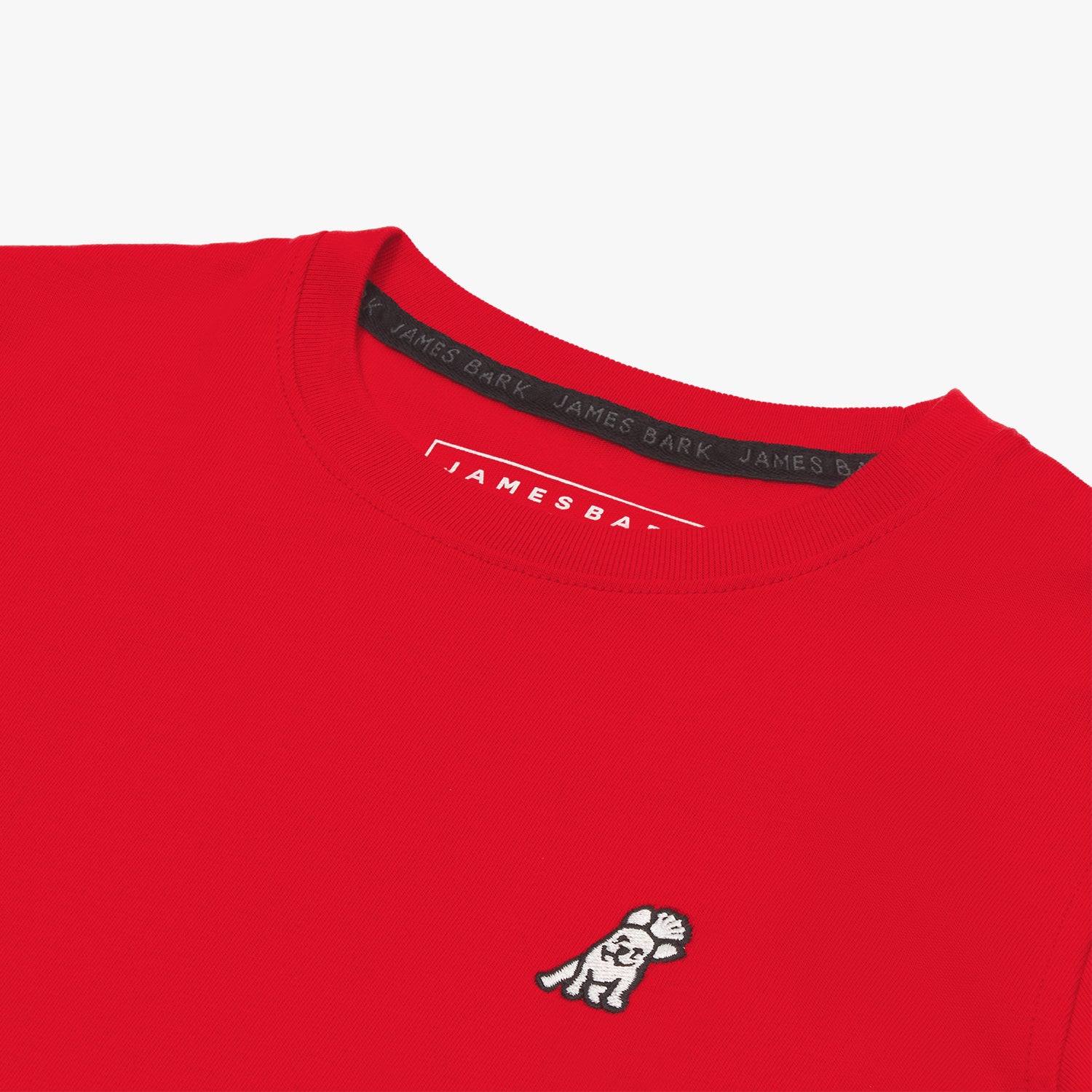 A close-up of the neckline and upper chest area of a red T-shirt, featuring a small embroidered white dog logo on the left side. The black inner collar lining displays the repeated 'JAMES BARK' branding, and the inside label reads 'JAMES BARK' in white text.