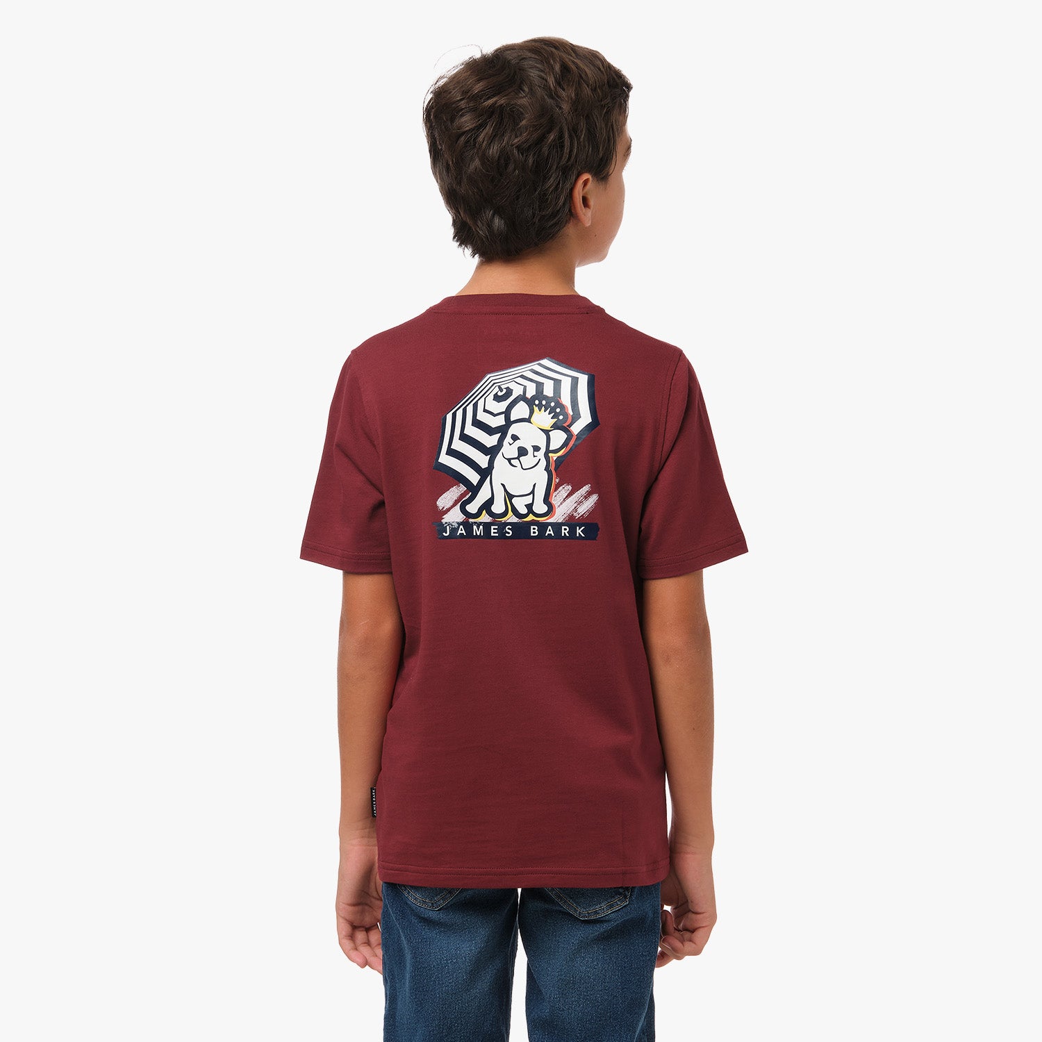 A young boy is shown from behind wearing a maroon T-shirt with a graphic on the back. The graphic features a dog sitting under a striped umbrella with the text "James Bark" below. He pairs the shirt with blue jeans.