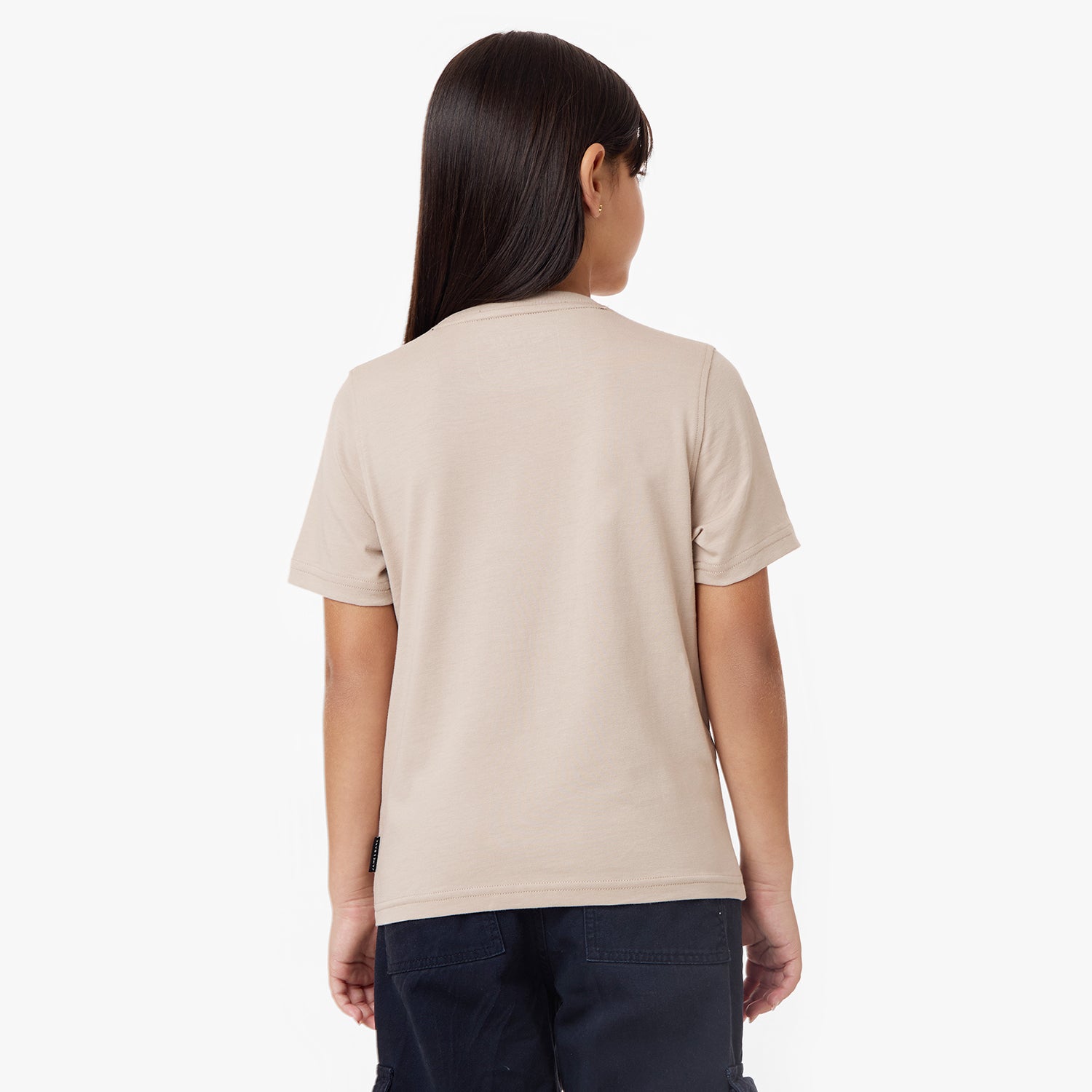 A young girl with long, sleek dark hair is shown from the back, wearing a beige short-sleeve t-shirt and navy cargo pants. The t-shirt has a minimal design with a small brand tag on the lower side seam. She stands against a plain white background with a relaxed posture.