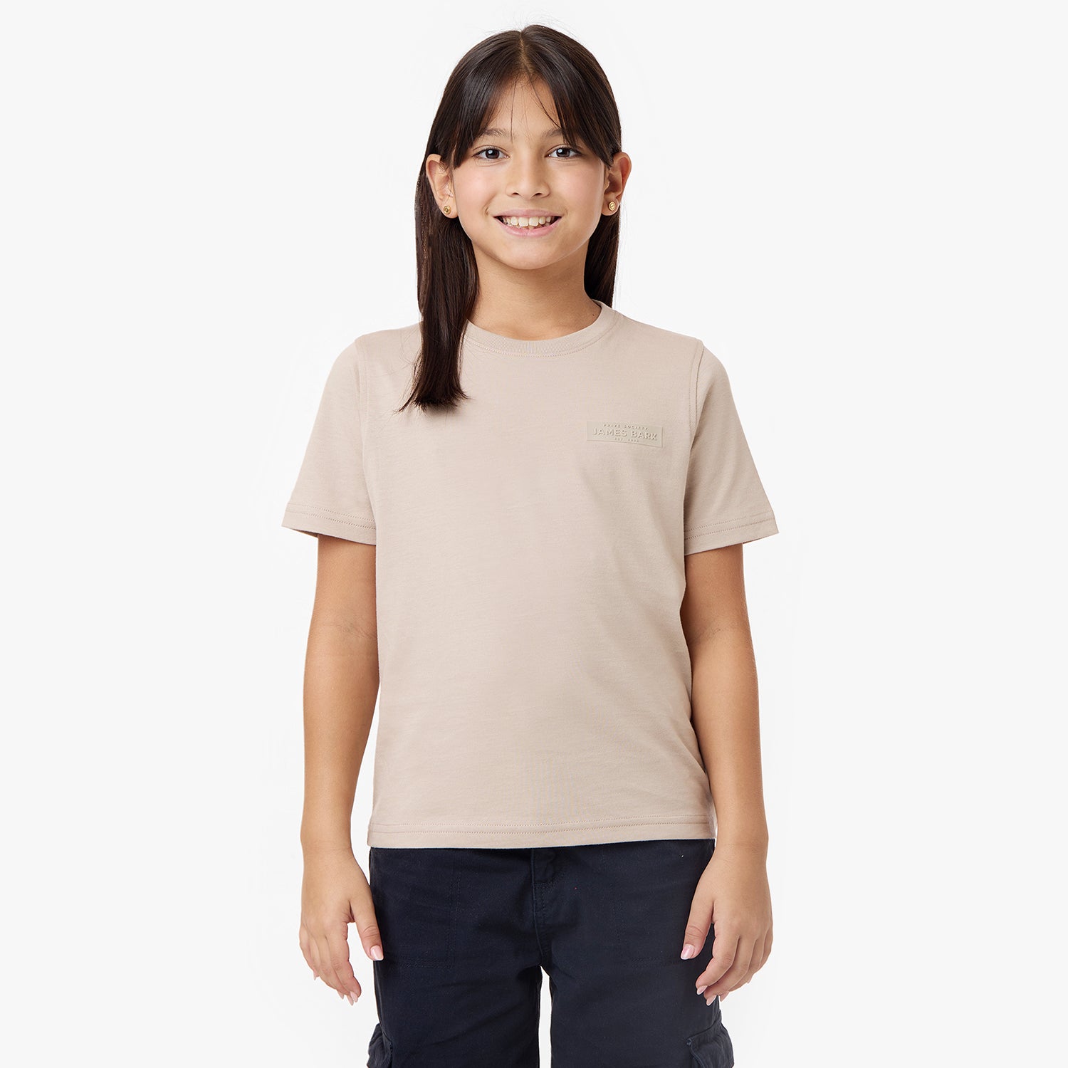 A young girl with long dark hair and small gold earrings smiles at the camera while standing against a plain white background. She is wearing a beige short-sleeve t-shirt with an embossed logo on the left chest and dark navy cargo pants. Her hands are relaxed at her sides, and she has a cheerful expression.