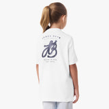 Back view of a young girl wearing a white T-shirt with a large decorative 'JB' logo and the text 'James Bark' on the back, paired with light pants