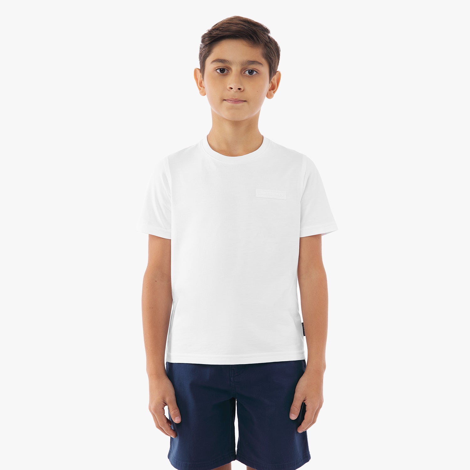 A young Boy with long dark hair and gold stud earrings wears a white t-shirt with a subtle embossed logo on the chest. She pairs it with light gray drawstring shorts featuring a small embroidered design. She stands against a plain white background with a neutral expression.