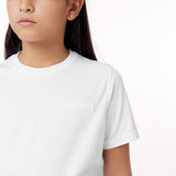 A close-up of a young girl wearing a white t-shirt with a minimalist embossed logo patch that reads "JAMES BARK" on the left side of her chest. The fabric is smooth, and the shirt has a classic round neckline and short sleeves. She has long dark hair and is wearing small gold stud earrings. The background is clean and white