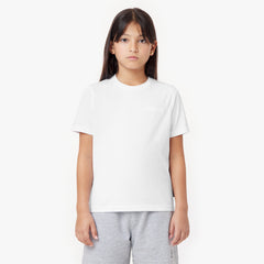 A young girl with long dark hair and gold stud earrings wears a white t-shirt with a subtle embossed logo on the chest. She pairs it with light gray drawstring shorts featuring a small embroidered design. She stands against a plain white background with a neutral expression.