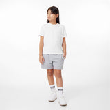 A young girl with long dark hair stands against a plain white background, wearing a casual white t-shirt with an embossed logo on the left chest and gray shorts with a small embroidered detail. She pairs her outfit with white sneakers, mid-length socks featuring navy blue stripes, and small gold earrings. She has a neutral expression and gazes off to the side with one hand in her pocket.