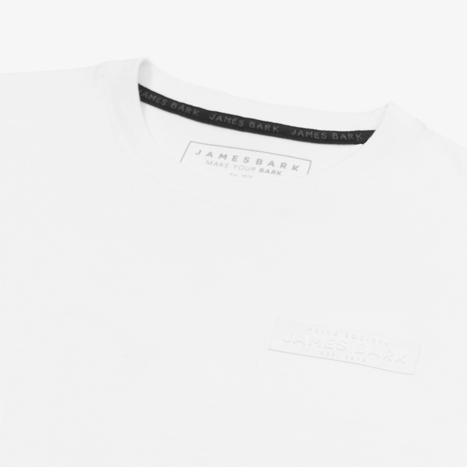 "A close-up of a white T-shirt, highlighting the tonal rubberized logo patch on the upper left chest that reads 'PRIVÉ SOCIETY JAMES BARK EST. 2015.' The neckline features a black inner lining with repeated 'JAMES BARK' branding, and the inside label displays 'JAMES BARK MAKE YOUR BARK' along with material details