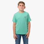 Front view of a boy wearing a mint green t-shirt with a small white embroidered logo on the chest, standing with his hands in his pockets.