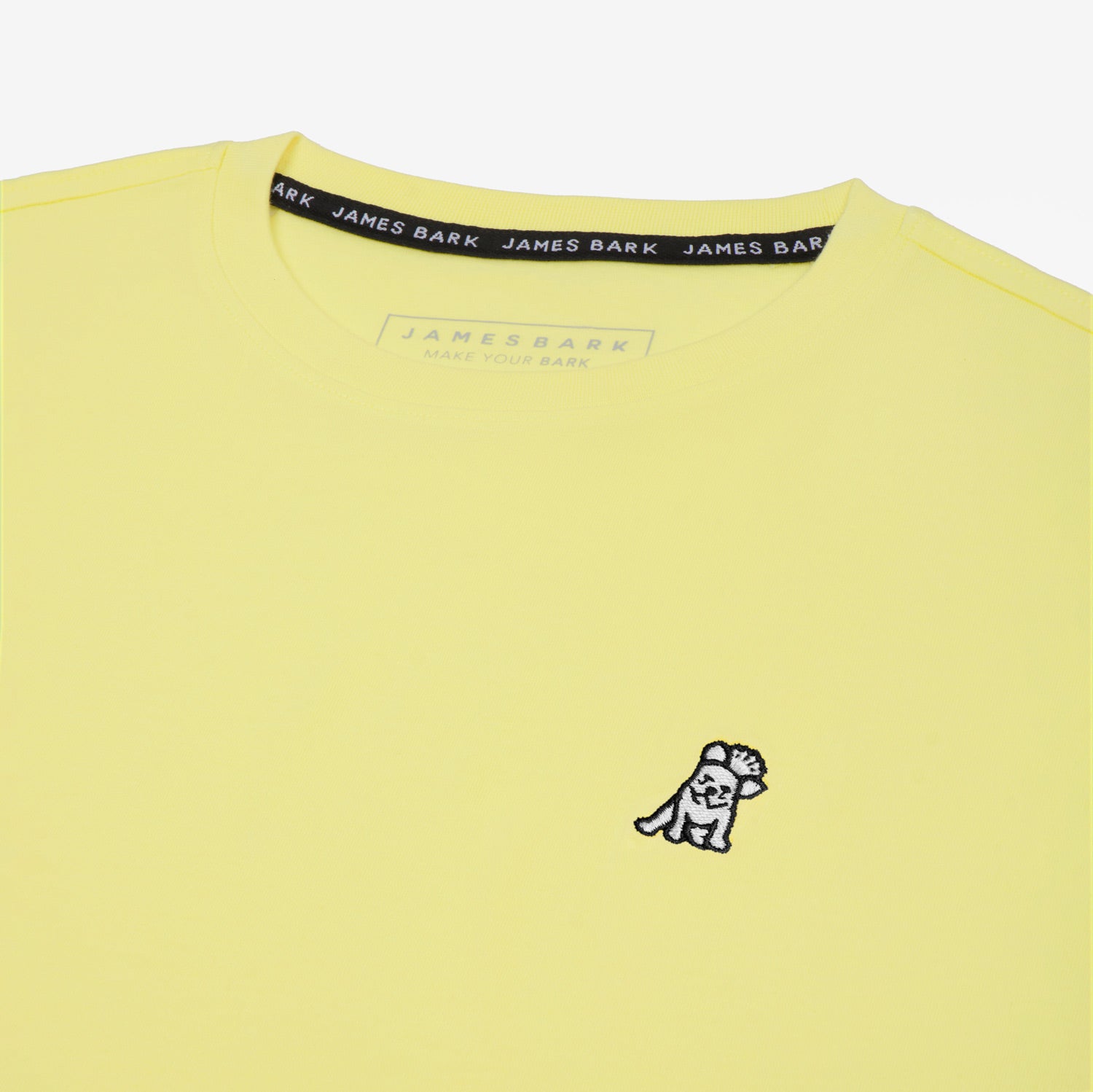 Close-up of the small white embroidered logo on the front of the light yellow t-shirt.