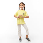 Full view of the young girl wearing the yellow T-shirt with white pants, standing with her hands by her sides.