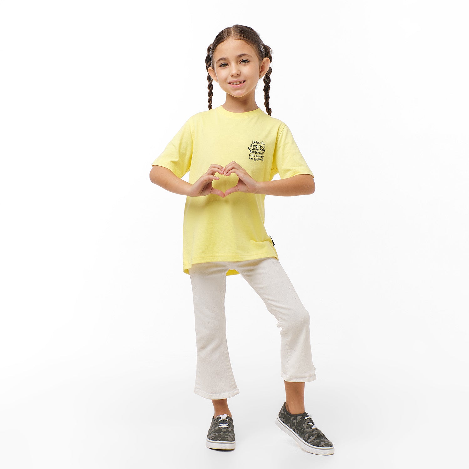 Full view of the young girl wearing the yellow T-shirt with white pants, standing with her hands by her sides.