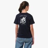 Back view of a woman wearing a navy blue T-shirt with a large decorative 'JB' logo and the text 'James Bark' on the back, paired with blue jeans.