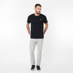 Full body shot of a man standing, wearing the black crew neck t-shirt with the white embroidered logo, paired with light grey trousers and black sneakers. He is looking off to his left with hands in his pockets