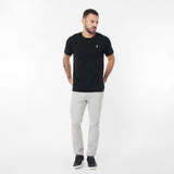 Full body shot of a man standing, wearing the black crew neck t-shirt with the white embroidered logo, paired with light grey trousers and black sneakers. He is looking off to his left with hands in his pockets