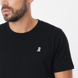 Close-up shot of the left chest area of the man wearing the black crew neck t-shirt, focusing on the ribbed neckline and the small white embroidered dog logo