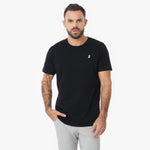 Front view waist-up shot of a man wearing the black crew neck t-shirt with the white embroidered logo, paired with light grey trousers. He is looking at the camera with one hand in his pocket