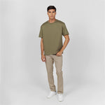 Full-body shot of a male model wearing an olive green oversized t-shirt with an embossed 'JAMES BARK ELEMENTS' logo on the chest. Paired with beige pants and white sneakers. Minimalist background.