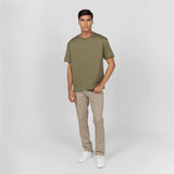 Full-body shot of a male model wearing an olive green oversized t-shirt with an embossed 'JAMES BARK ELEMENTS' logo on the chest. Paired with beige pants and white sneakers. Minimalist background.