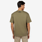 Back view of a male model wearing an olive green oversized t-shirt. Features a minimalist design with a black rectangular 'JAMES BARK' label near the neckline. Paired with beige pants. White background