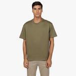 Male model wearing an olive green oversized t-shirt with a minimalist design. Features a subtle embossed 'JAMES BARK' logo on the chest and a ribbed crew neckline. Paired with beige pants. White background