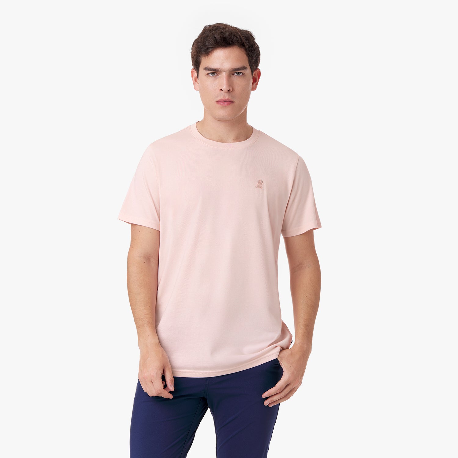 Male model wearing a light pink short-sleeved t-shirt with a matching embroidered dog logo and navy pants, standing front view with one hand in pocket