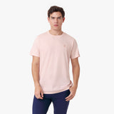 Male model wearing a light pink short-sleeved t-shirt with a matching embroidered dog logo and navy pants, standing front view with one hand in pocket