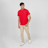 A male model wearing a red short-sleeve t-shirt with a small embroidered dog logo on the left chest, paired with beige slim-fit pants and white sneakers with red accents. He is posing in a relaxed stance against a neutral gray background, looking off to the side. The outfit is casual yet polished, with a clean and minimal aesthetic.