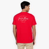 A man seen from behind wearing a vibrant red short-sleeve t-shirt with white printed text on the upper back that reads: "PRIVÉ SOCIETY, James Bark, make your bark, BASED IN USA, EST. 2015." He is also wearing beige pants. The background is a clean white backdrop, highlighting the contrast of the t-shirt's bold color and the crisp white typography